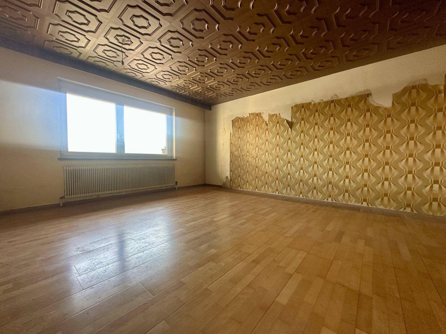 Geräumiges Wohnzimmer mit Parkettboden, gemusterter Decke und Tapetenresten.