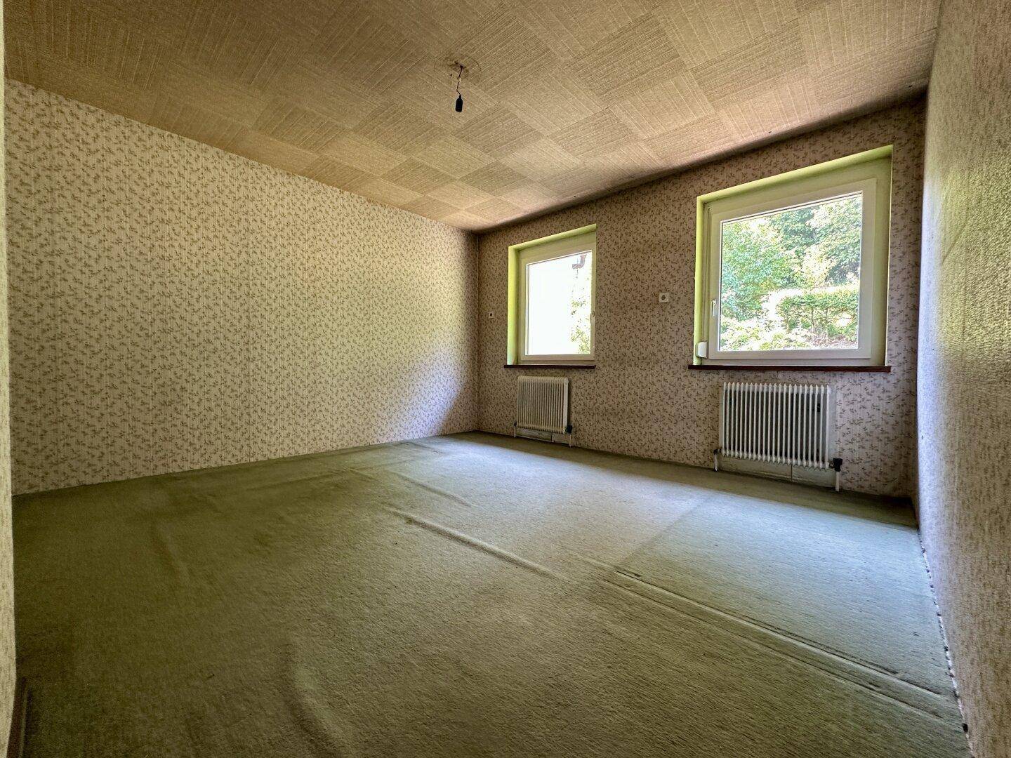 Schlafzimmer mit grünem Teppichboden, gemusterter Tapete und zwei Fenstern.