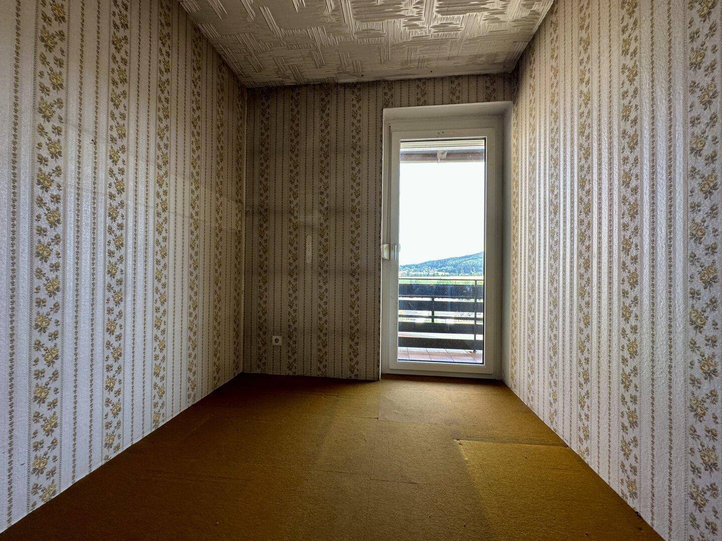 Kleines Zimmer mit gemusterter Tapete, Teppichboden und Balkonzugang.
