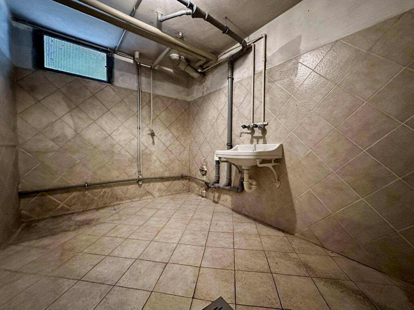 Kellerraum mit Waschbecken, Fliesenwänden und sichtbaren Rohren.