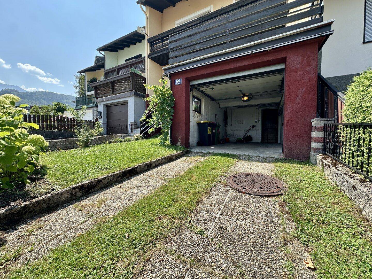 Vorgarten mit Garage, Auffahrt und blühenden Pflanzen an einem sonnigen Tag.