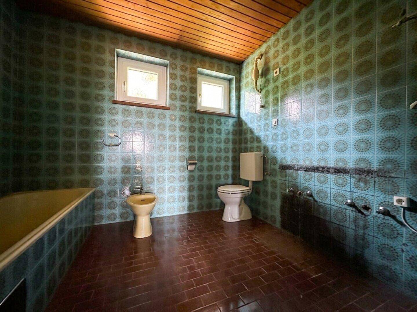 Badezimmer im Stil der 80er Jahre mit blauen Fliesen, Badewanne, Bidet und zwei Fenstern.