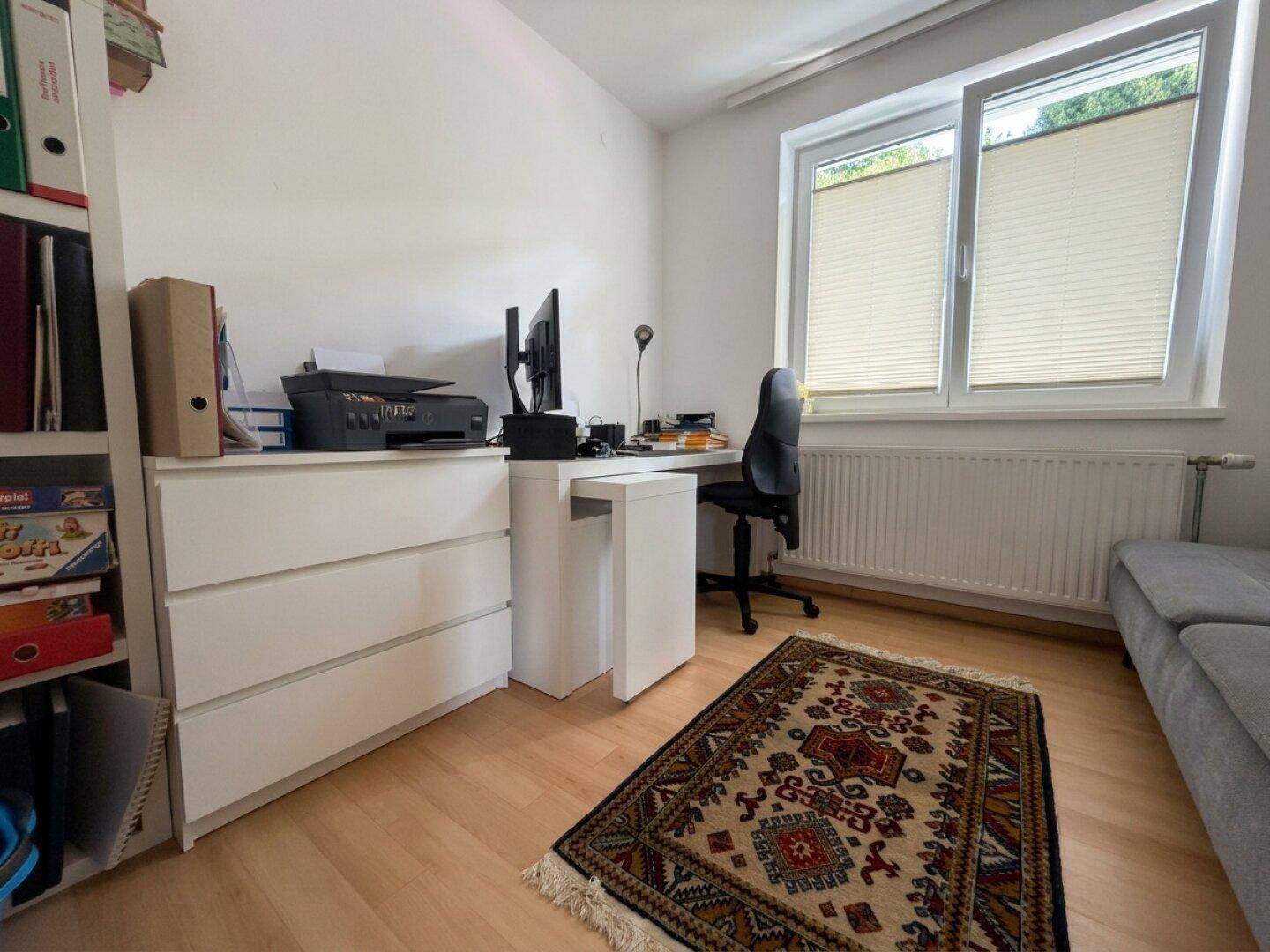 Helles Arbeitszimmer mit Schreibtisch, Bürostuhl, weißen Schubladen und Fenster mit Jalousien.