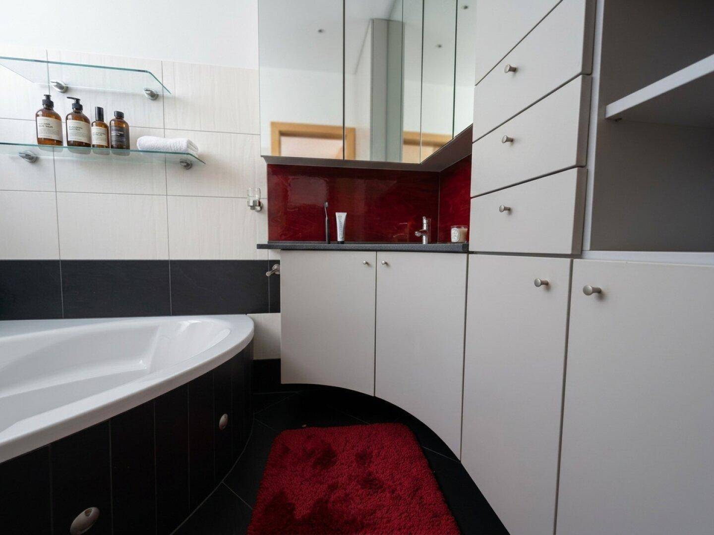 Badezimmer mit Eckbadewanne, schwarz-weißen Fliesen und weißen Schränken mit roter Akzentwand.