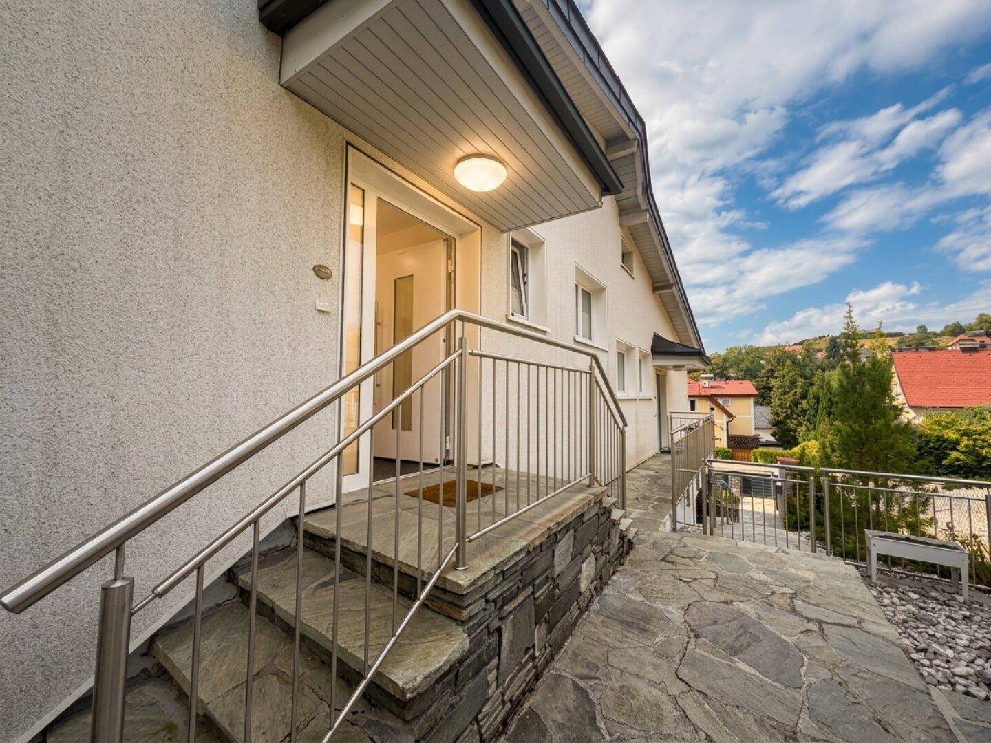 Eingangsbereich des Hauses mit Steinstufen, Edelstahlgeländer und moderner Haustür.