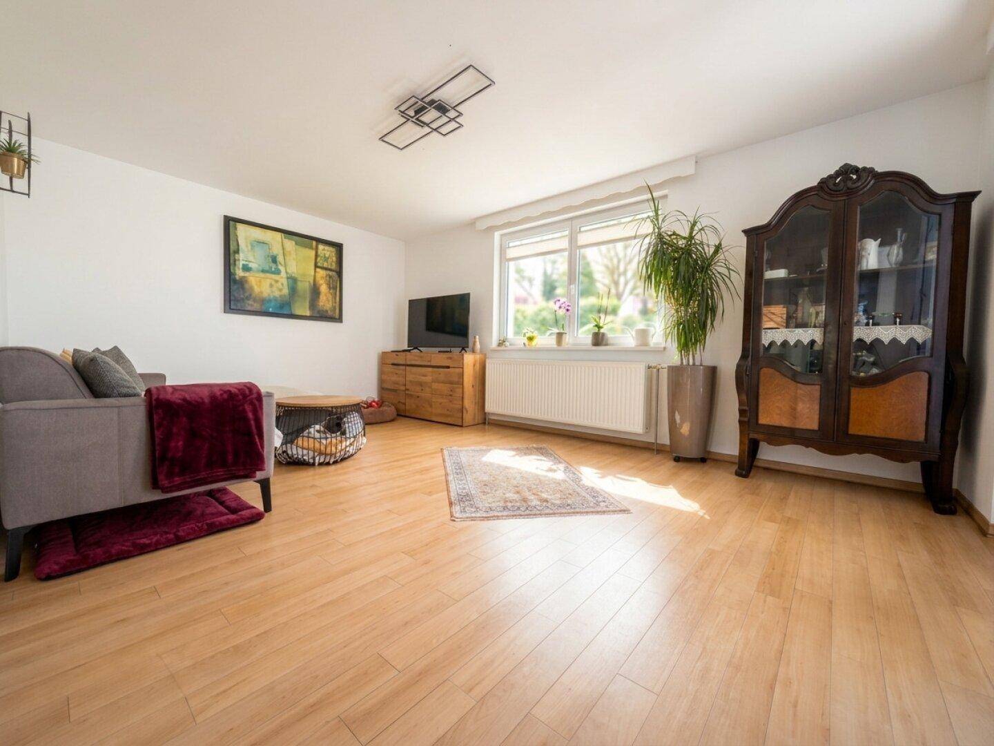 Helles Wohnzimmer mit hellem Holzboden, grauem Sofa, Fernseher und traditioneller Vitrine.