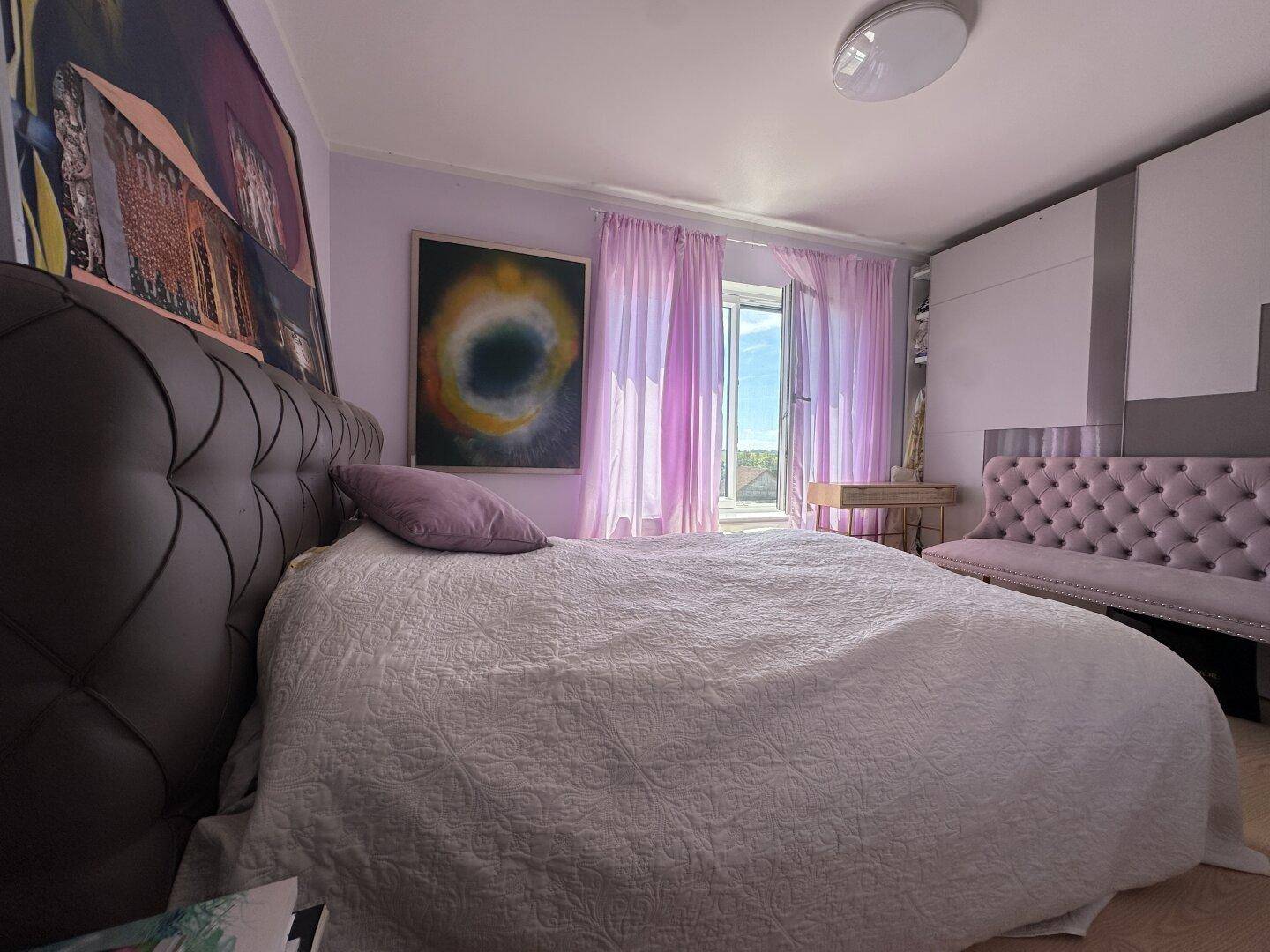 Schlafzimmer mit lila Vorhängen, gepolstertem Bett und dekorativen Kunstwerken an den Wänden.