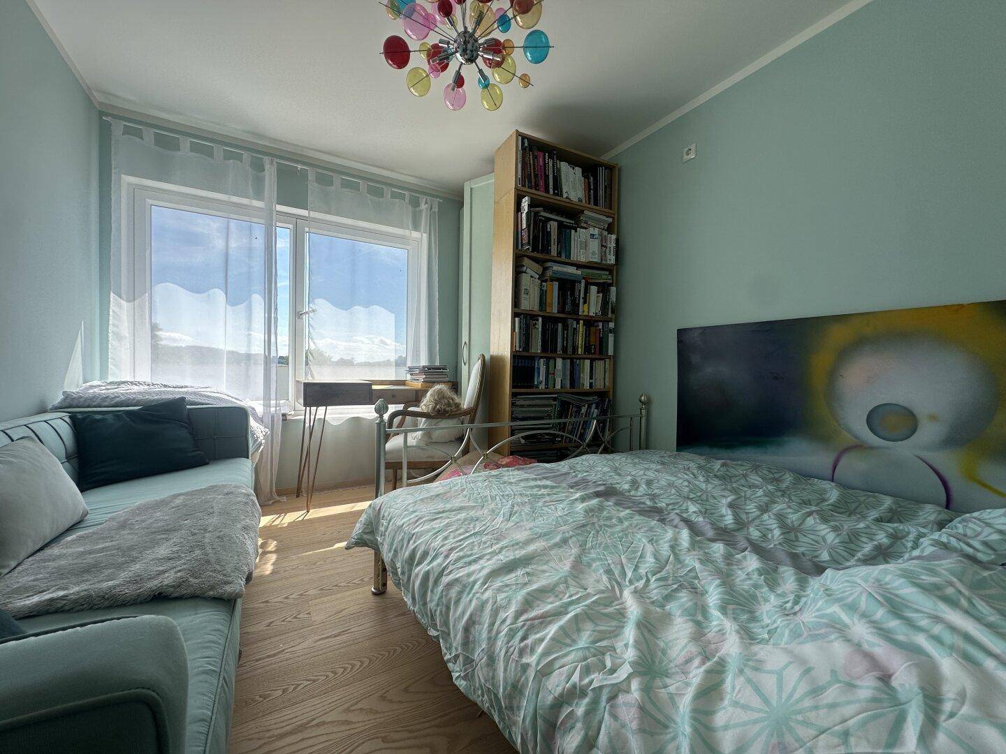 Helles Schlafzimmer mit mintgrünen Wänden, Holzboden und einem Bücherregal am Fenster.