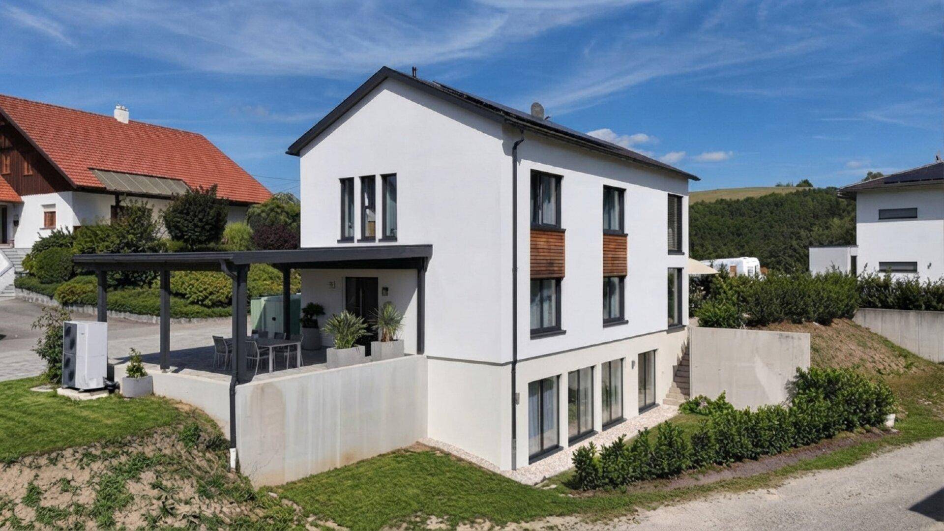 Modernes Einfamilienhaus mit weißer Fassade, dunklen Fensterrahmen und Carport in ländlicher Umgebung.