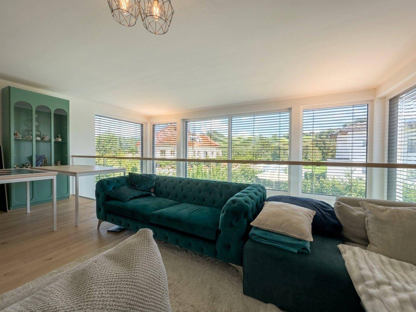 Helles Wohnzimmer mit großen Fenstern, grüner Couch und Blick auf die grüne Umgebung.