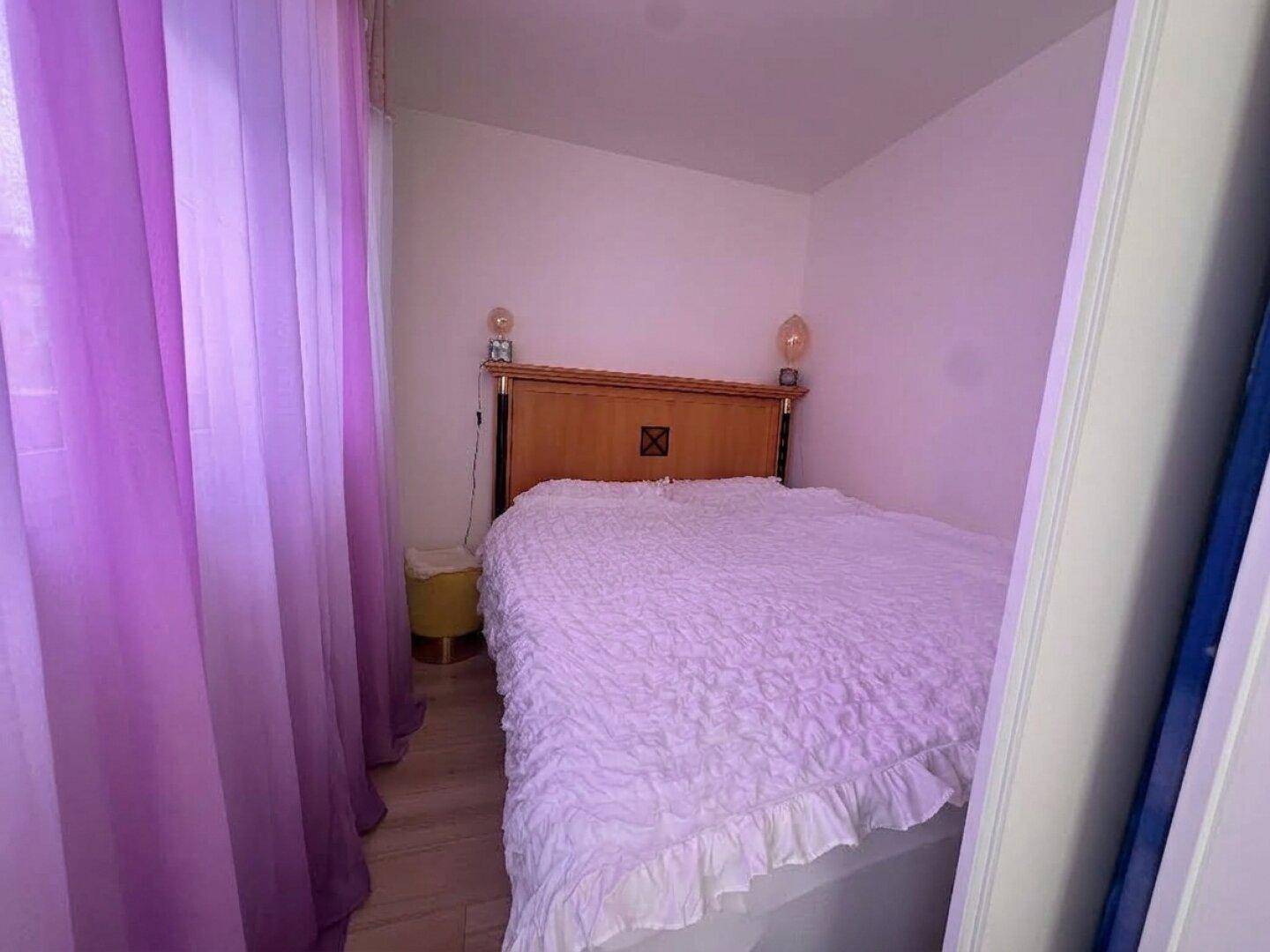Kleines Schlafzimmer mit Doppelbett, Holzboden und lila Vorhängen, hell beleuchtet.