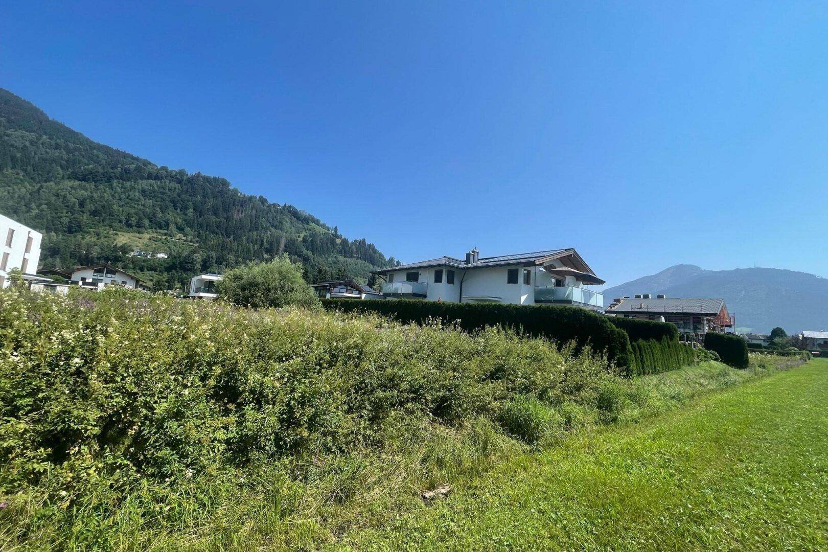Modernes Haus mit Balkonen und gepflegter Hecke, eingebettet in eine grüne Landschaft mit Bergblick unter strahlend blauem Himmel.