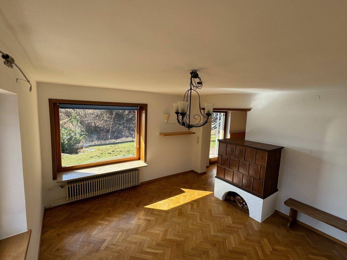 Helles Wohnzimmer mit traditionellem Kachelofen, Parkettboden und Blick ins Grüne.