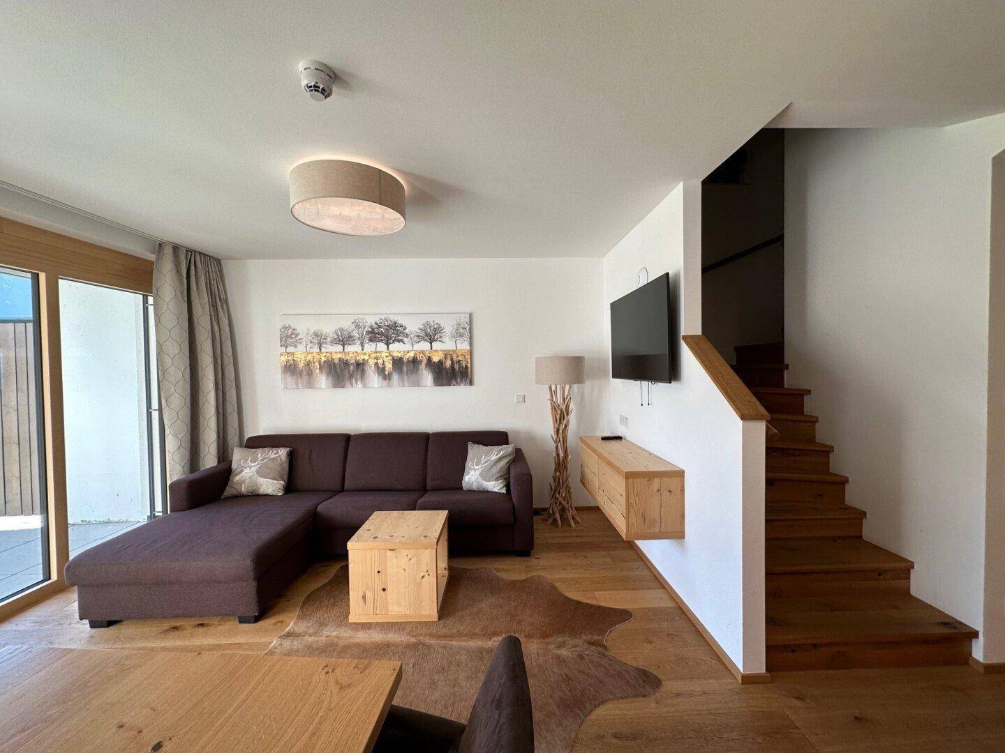 Heller Wohnbereich mit bequemer Couch, Holzboden und offener Holztreppe zum Obergeschoss.