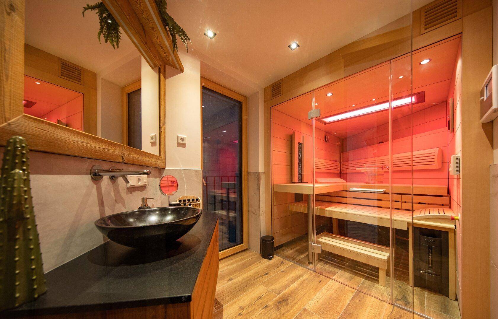 Modernes Badezimmer mit Waschbecken, Spiegel und einer verglasten Sauna mit stimmungsvoller Beleuchtung.