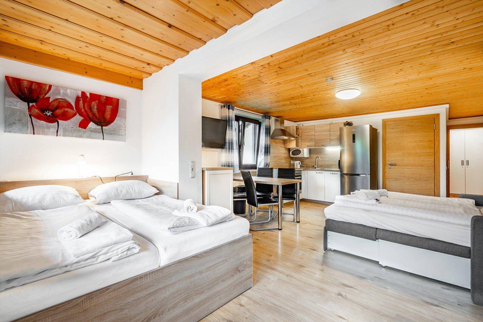 Helles Studio-Apartment mit Doppelbett, kleiner Küchenzeile und Essbereich unter Holzbalkendecke.