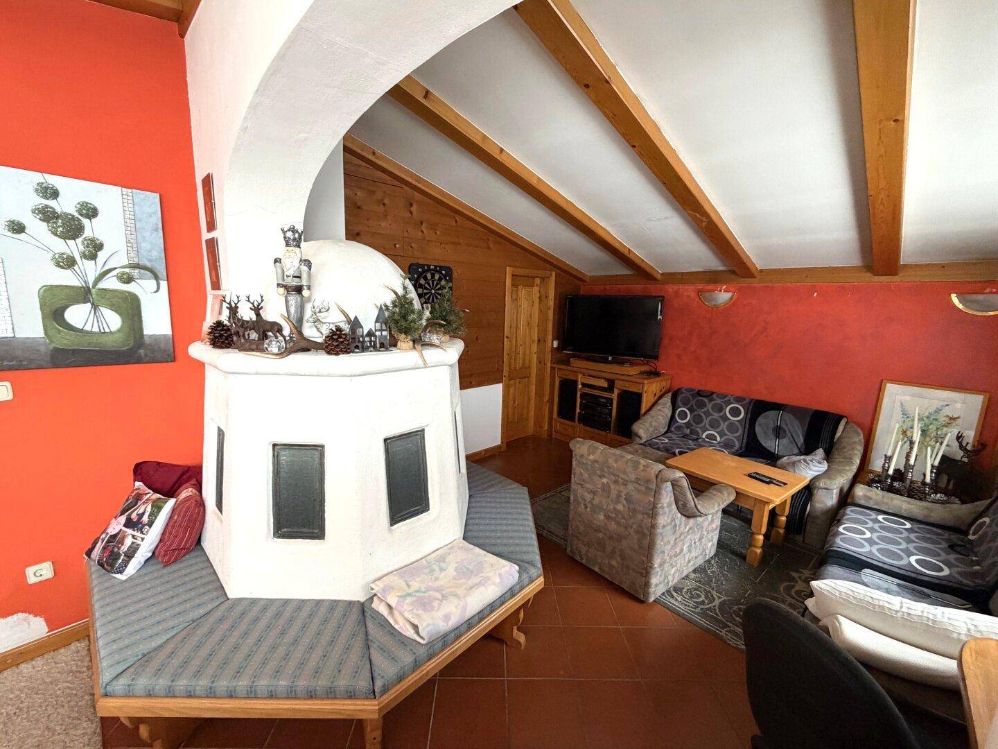 Gemütliches Wohnzimmer mit traditionellem Kachelofen, roten Wänden und sichtbaren Holzbalken.