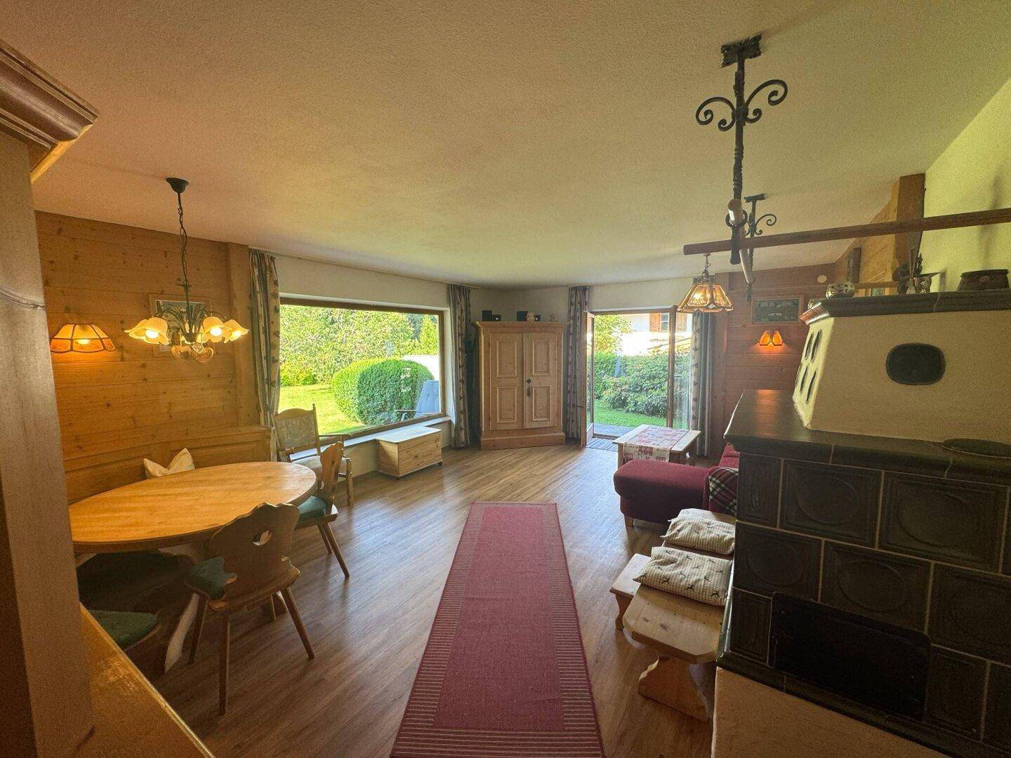 Rustikaler Wohn-Essbereich mit Holzwänden, Kachelofen und großem Fenster zum Garten.