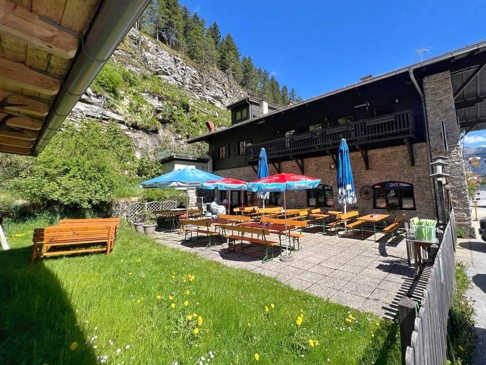 Gemütlicher Außenbereich mit Biertischen, Sonnenschirmen und einer Rasenfläche vor dem traditionellen Gebäude.