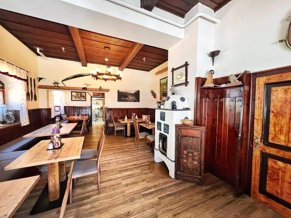 Rustikaler Speisesaal mit Holztischen, Bänken, einem Kachelofen und einer Holzdecke mit sichtbaren Balken.