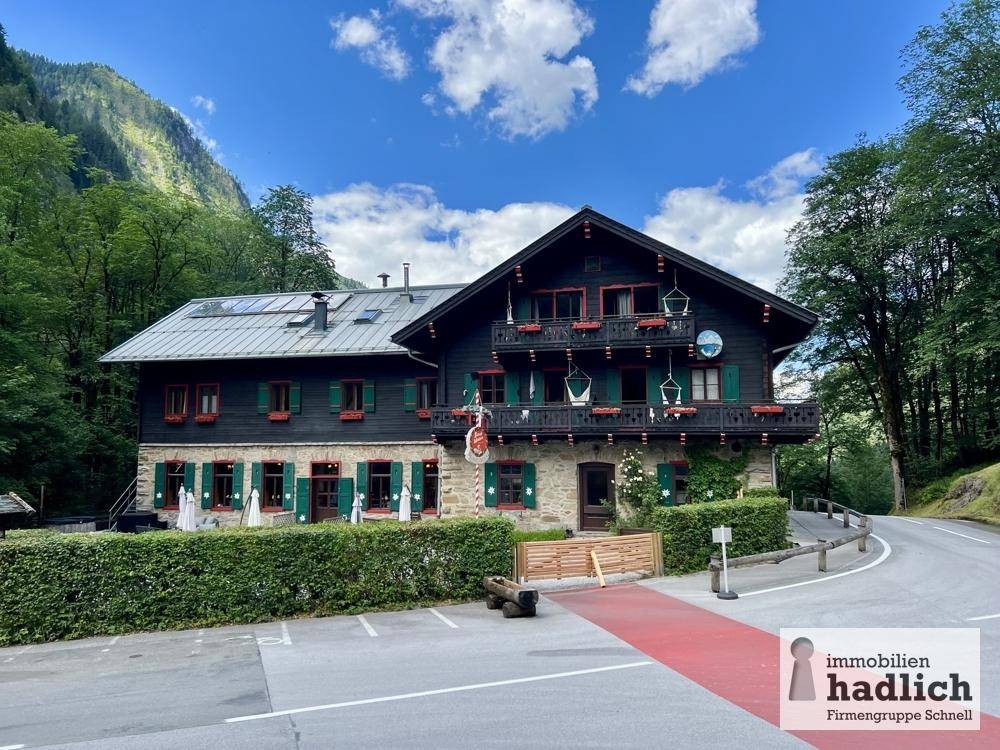 Traditionelles Gebäude mit dunklem Holz und Stein, umgeben von Bergen und Bäumen, mit Parkplatz.