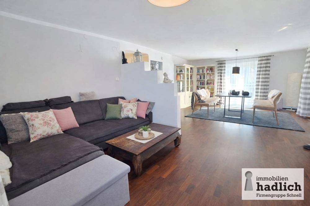 Gemütlicher Wohn-Essbereich mit dunklem Sofa, Holzboden und viel Tageslicht.