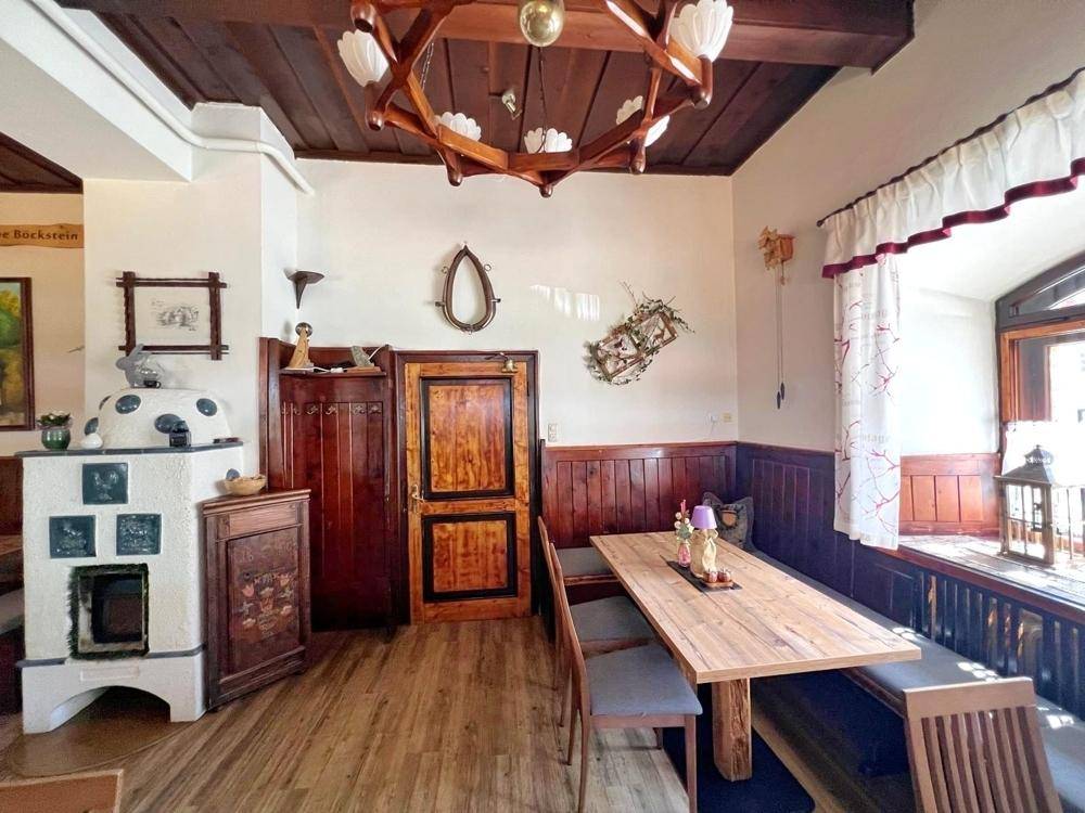 Gemütlicher Essbereich mit Holztischen, Bänken, einem traditionellen Kachelofen und einem Fenster mit Vorhängen.