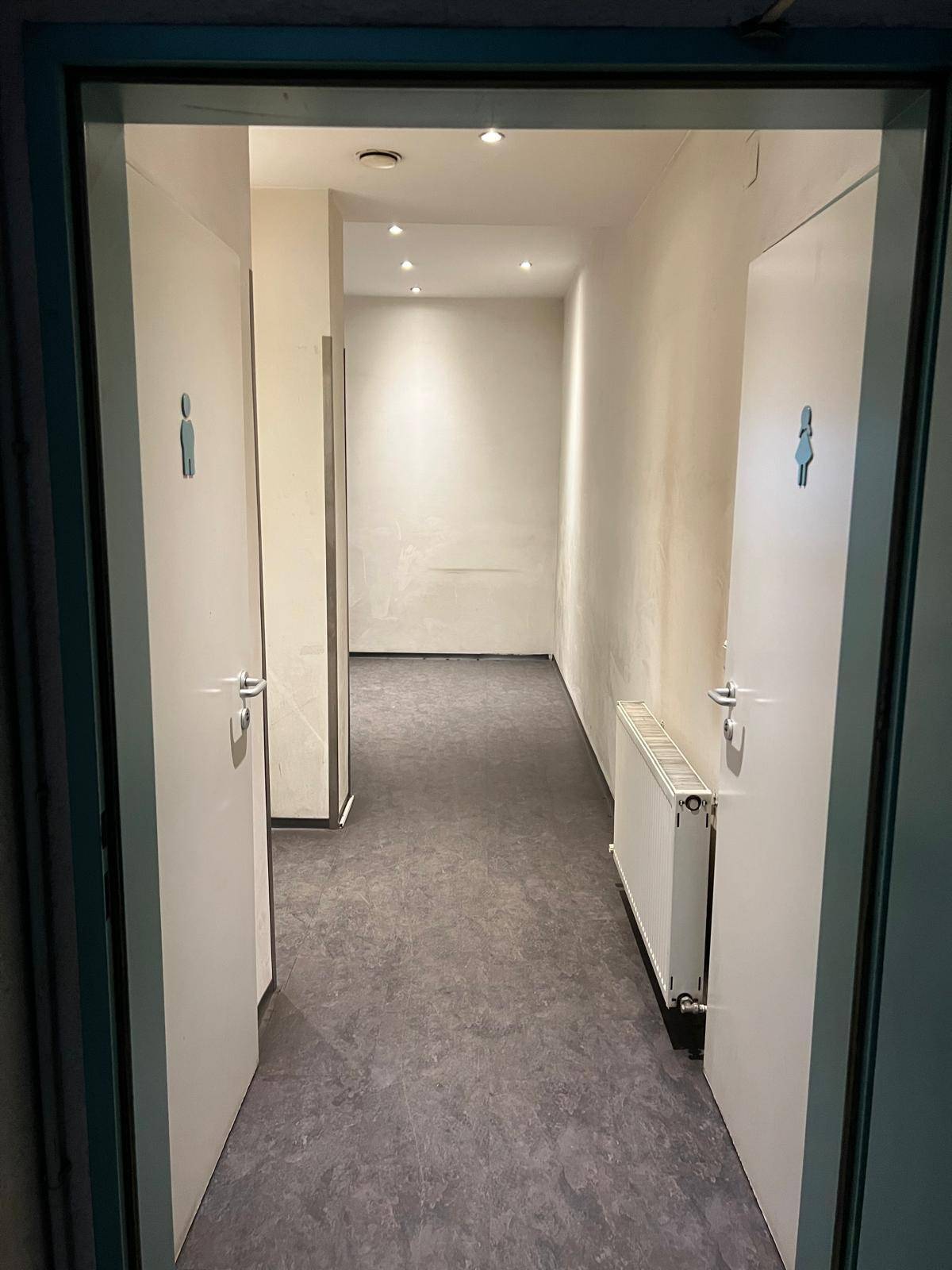 Flur mit dunklem Boden und zwei Türen, die zu getrennten Toilettenräumen führen, mit einem Heizkörper.