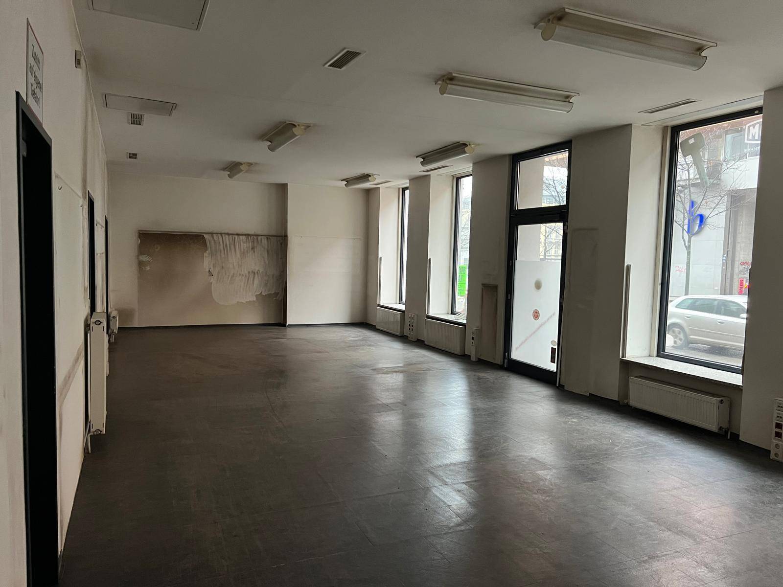 Großer, offener Geschäftsraum mit vielen Fenstern zur Straße und dunklem Boden, der renoviert werden muss.