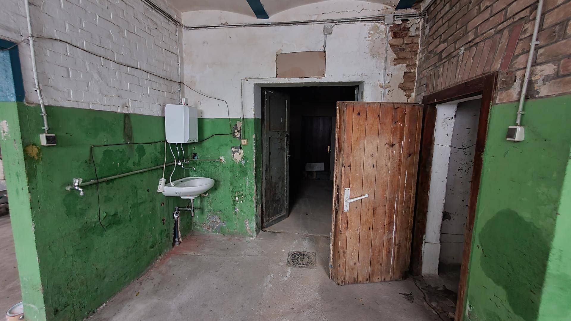 Kellerraum mit Waschbecken, abgenutzten Wänden, grüner Farbe und mehreren Holztüren in schlechtem Zustand.