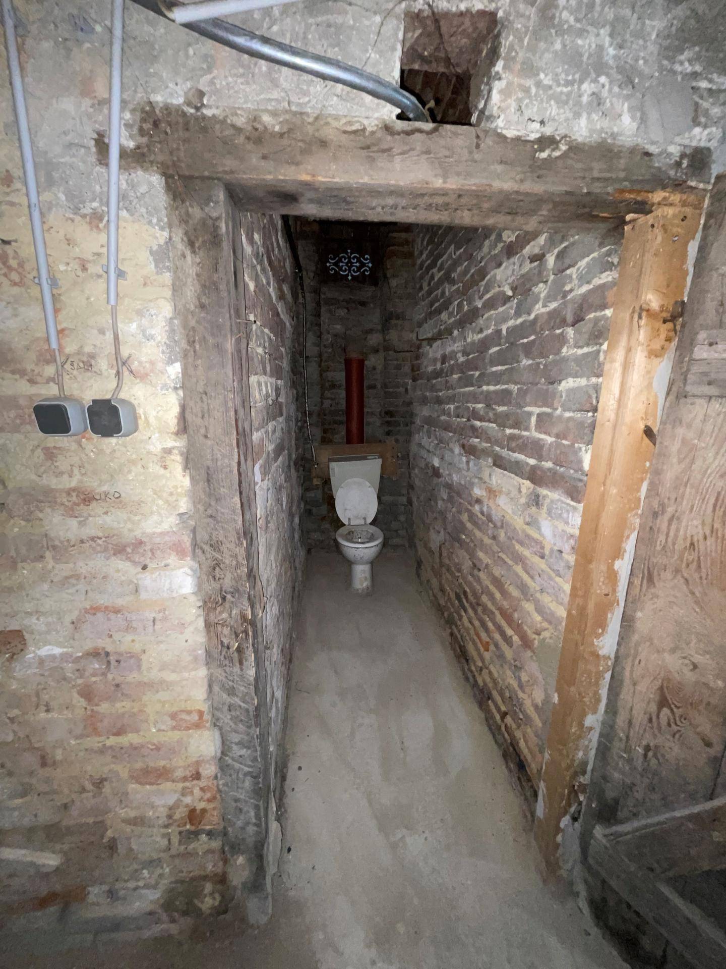 Schmale Toilettenkabine im Keller mit unverputzten Ziegelwänden und einfacher Toilette.