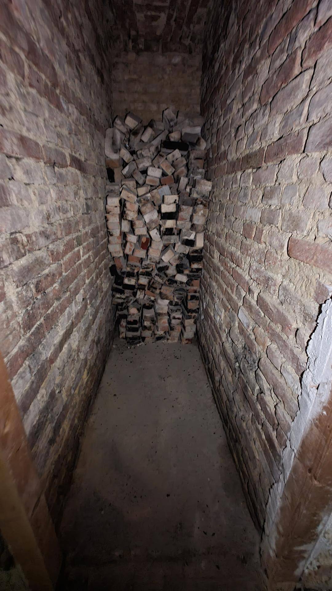 Schmaler Kellerraum mit unverputzten Ziegelwänden und einem Stapel Ziegelsteine am Ende des Raumes.