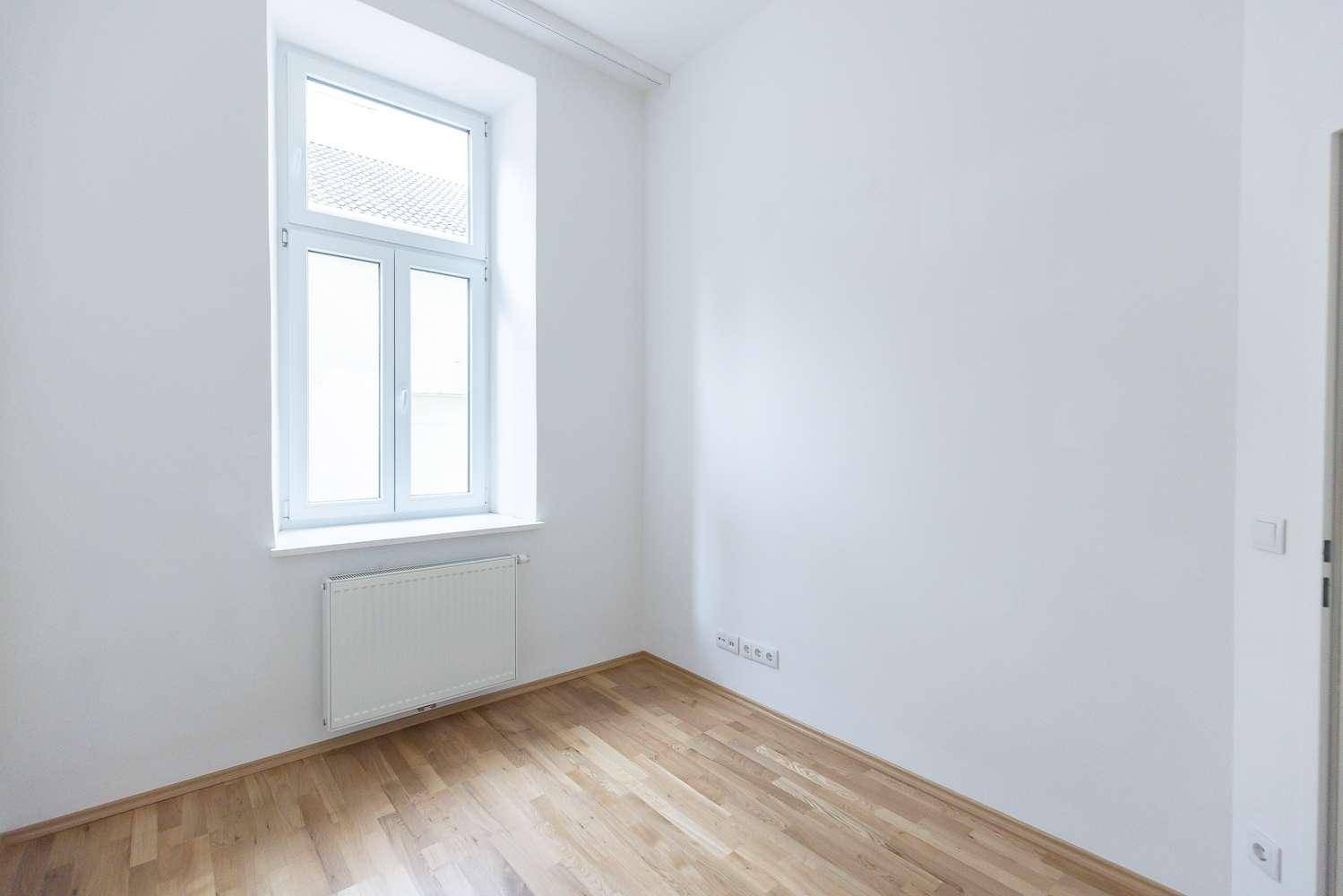 Leeres Zimmer mit hellem Parkettboden und einem großen Fenster, das viel Tageslicht hereinlässt.