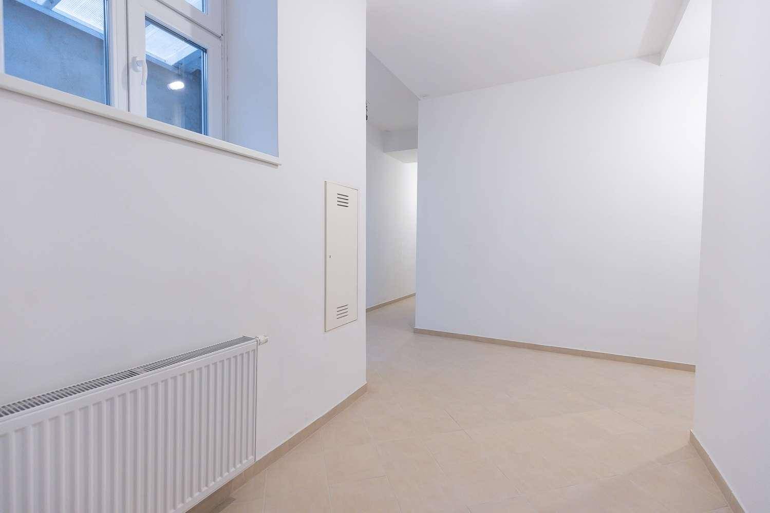 Langer, heller Flur mit weißen Wänden, Fliesenboden und einem Fenster, das Tageslicht hereinlässt.