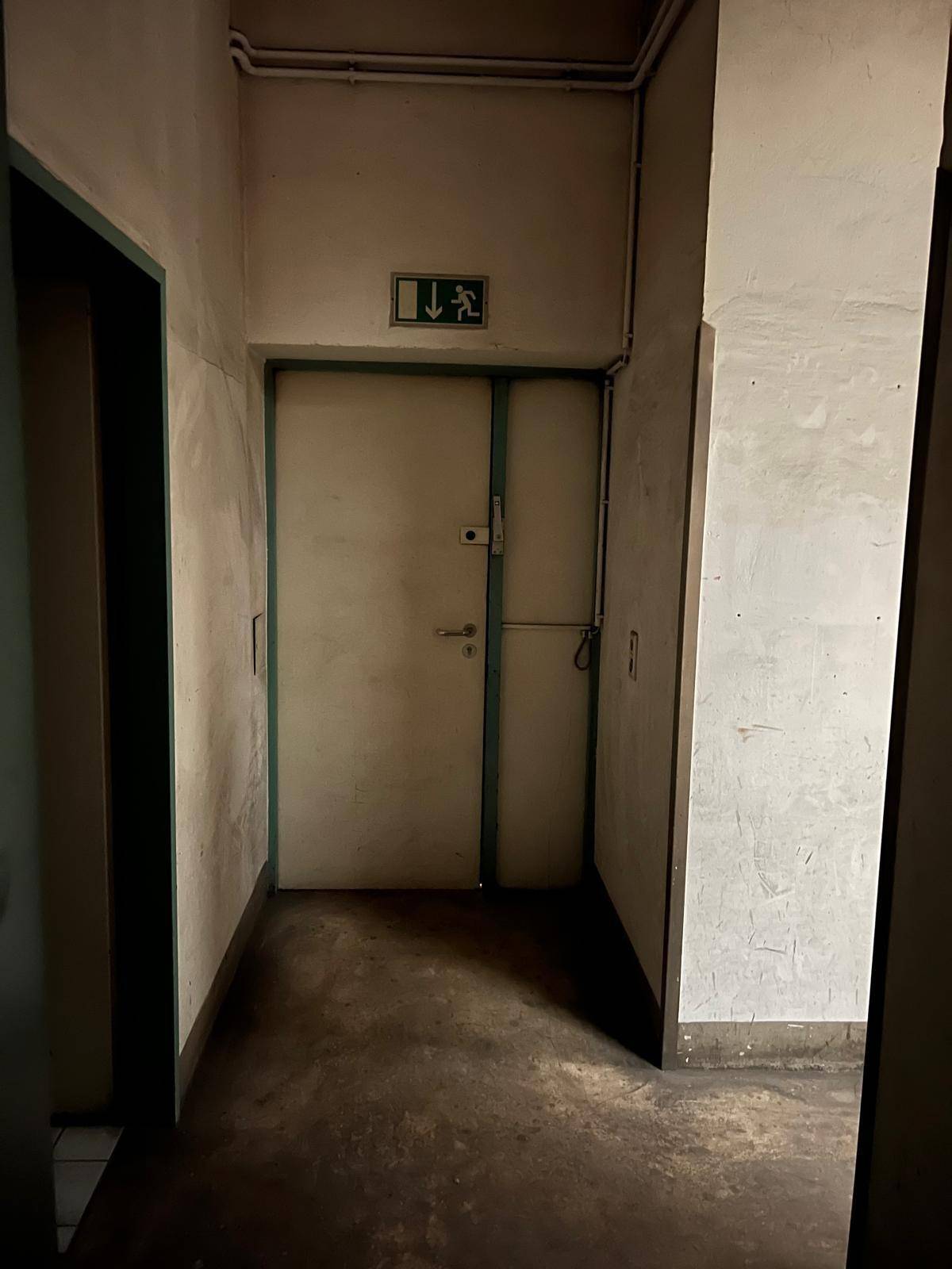 Schmaler Flur mit einer alten Tür und einem Notausgangsschild, der renovierungsbedürftig ist.