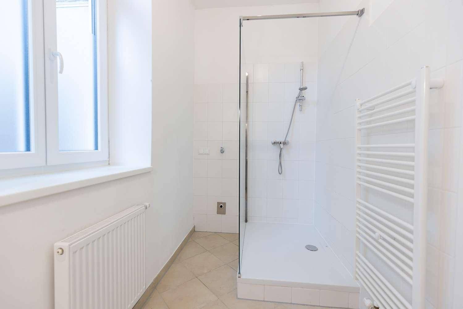 Helles Badezimmer mit Duschkabine, weißen Wandfliesen und einem Heizkörper unter dem Fenster.