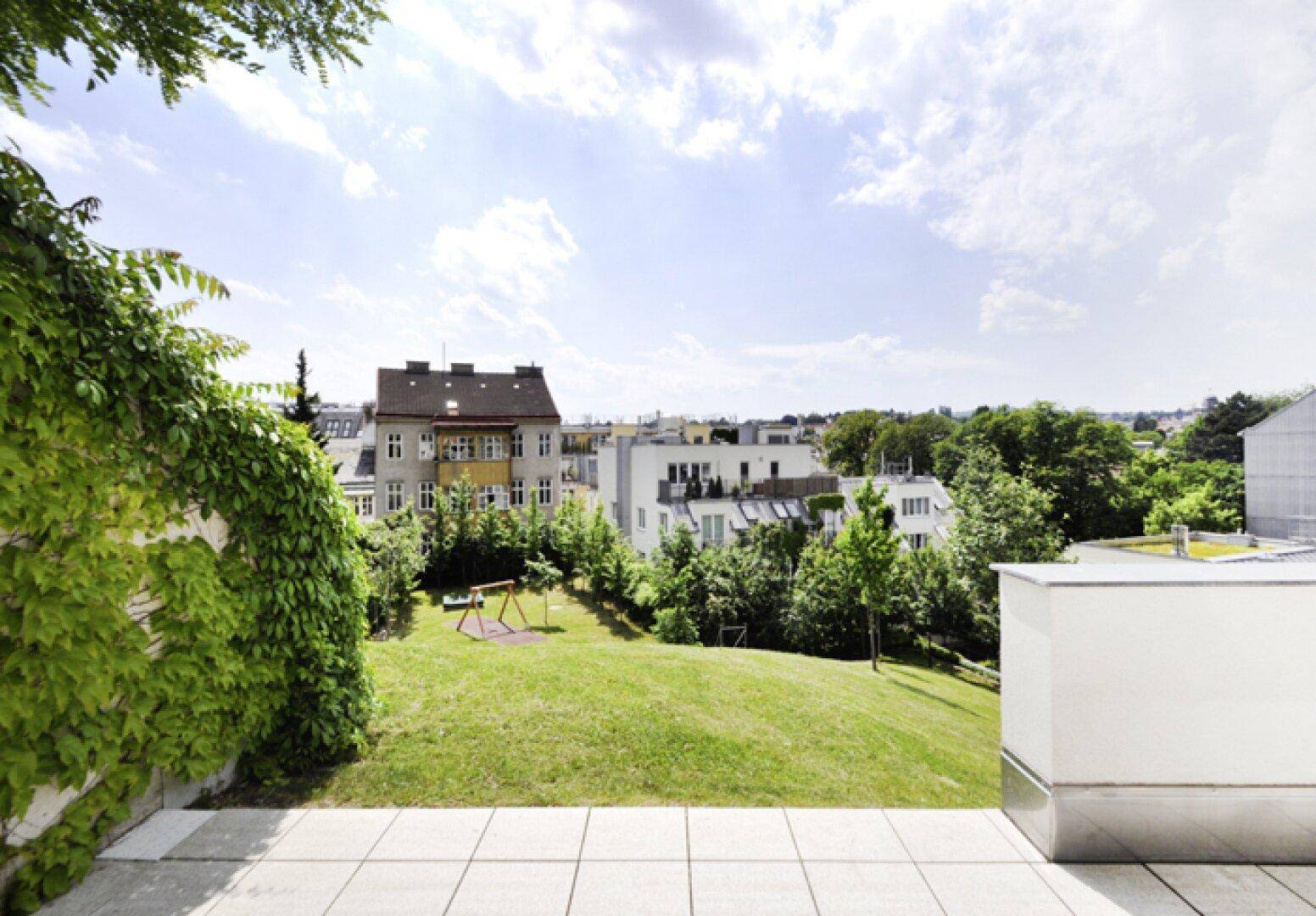 Blick von der Terrasse auf einen großen Rasenbereich mit Spielplatz und umliegenden Gebäuden.