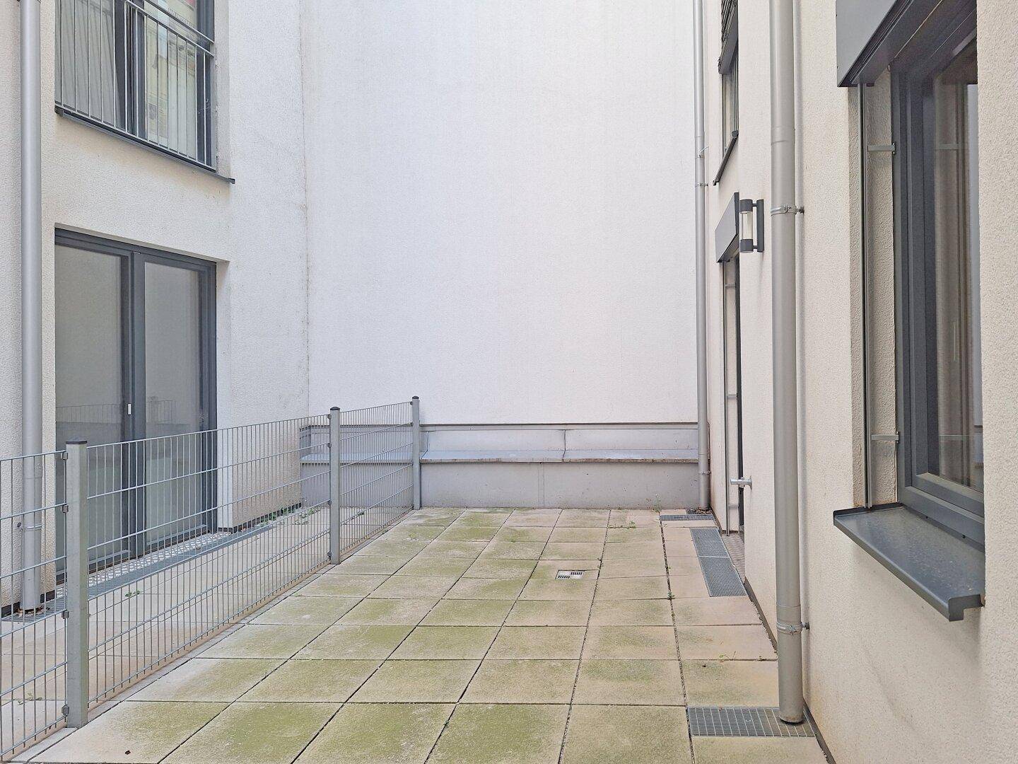 Kleine gepflasterte Terrasse im Innenhof mit Metallzaun und Zugang zum Gebäude.