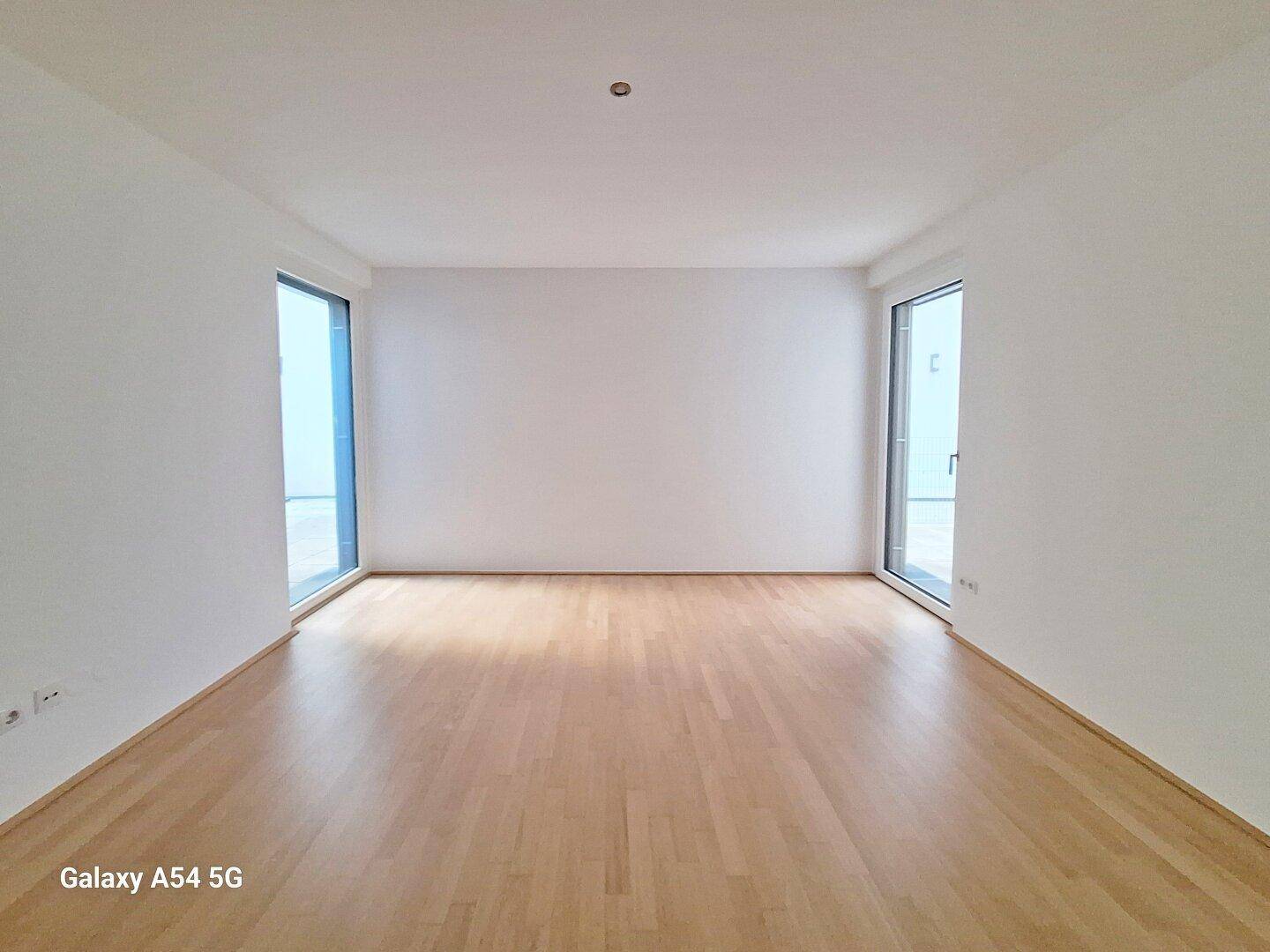 Heller, leerer Raum mit Parkettboden und zwei hohen Fenstern.