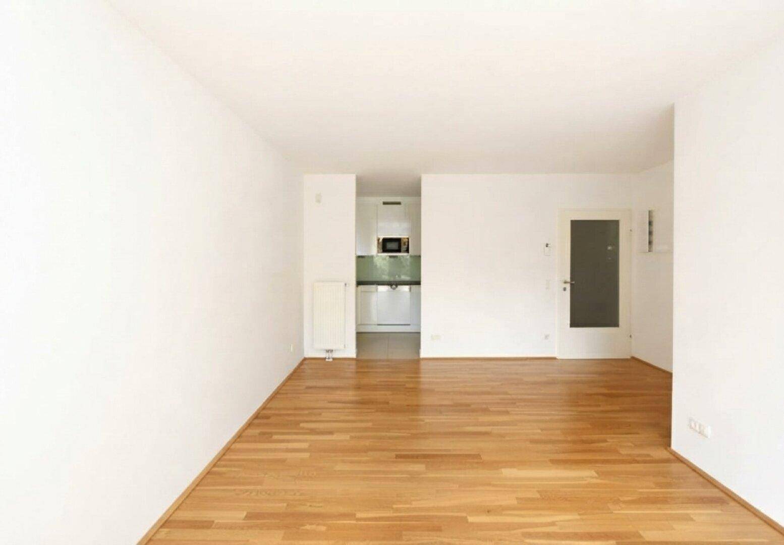 Geräumiges, unmöbliertes Wohnzimmer mit hellem Parkettboden und Zugang zur Küche.