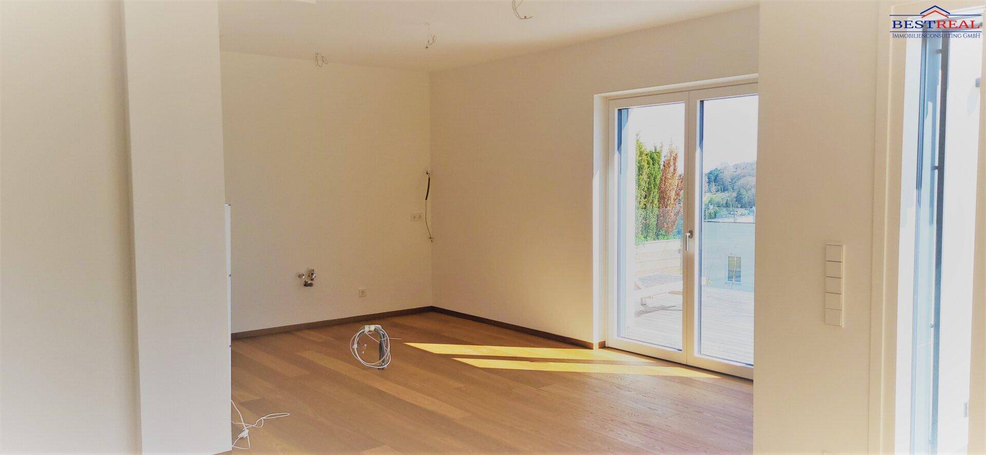 Leerer Raum mit Holzboden und großem Fenster, das viel Tageslicht hereinlässt und Blick ins Grüne bietet.