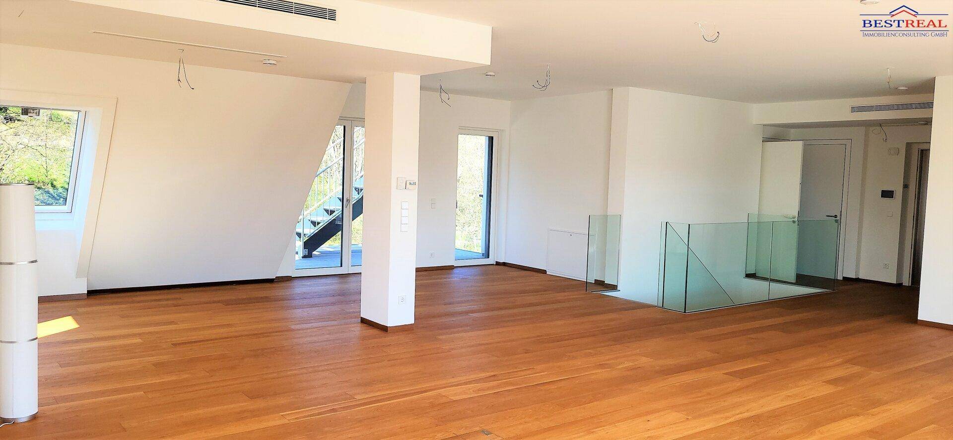 Geräumiger, heller Wohnbereich mit Holzfußboden und moderner Treppe mit Glasgeländer.