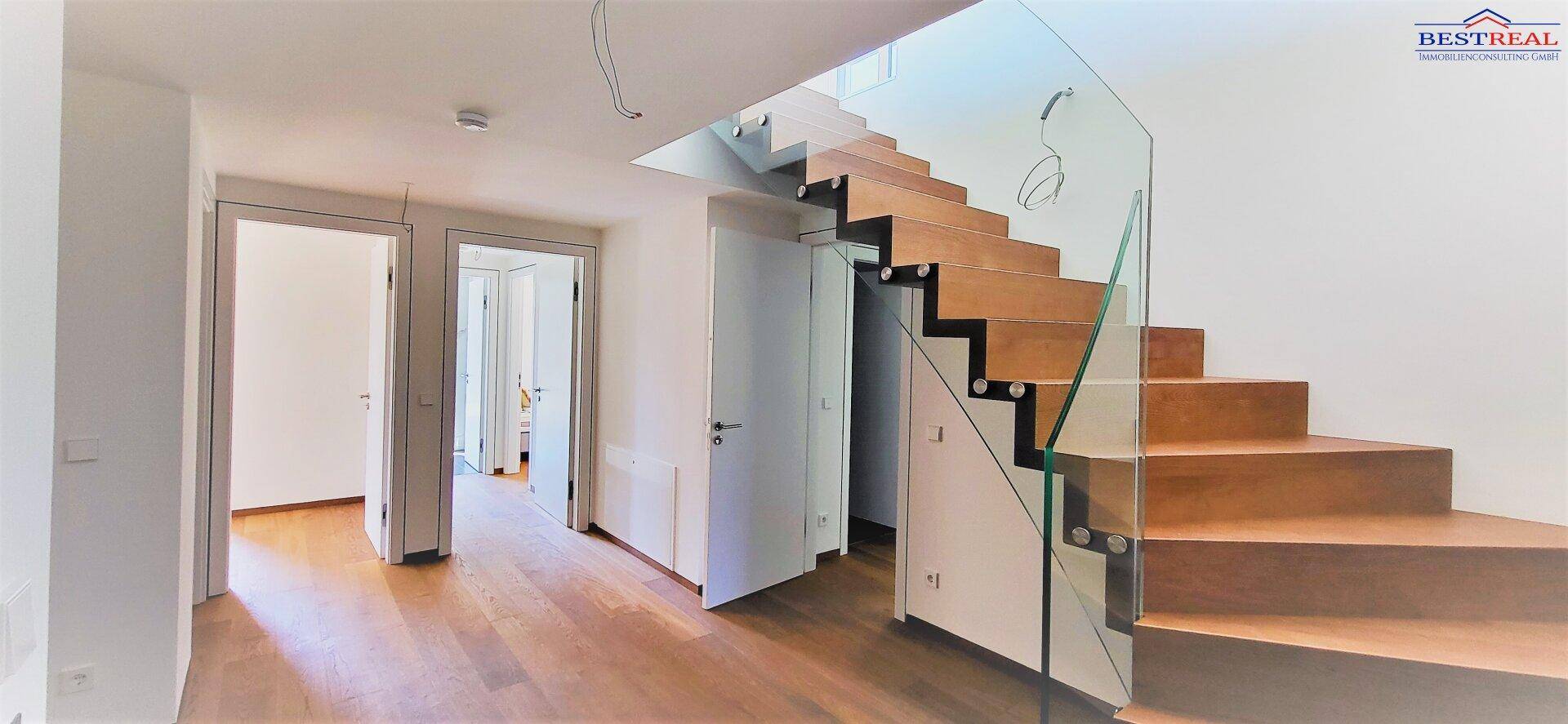 Heller Flur mit moderner Holztreppe und Glasgeländer, der zu weiteren Räumen führt.
