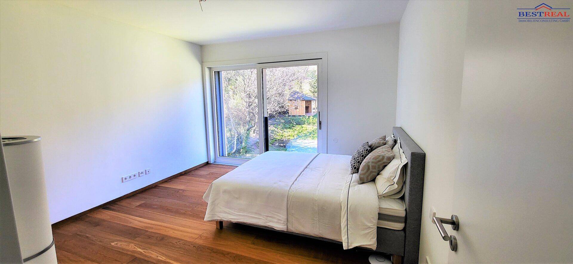 Schlafzimmer mit Holzboden, Bett und großem Fenster mit Zugang zu einem Balkon und Blick ins Grüne.