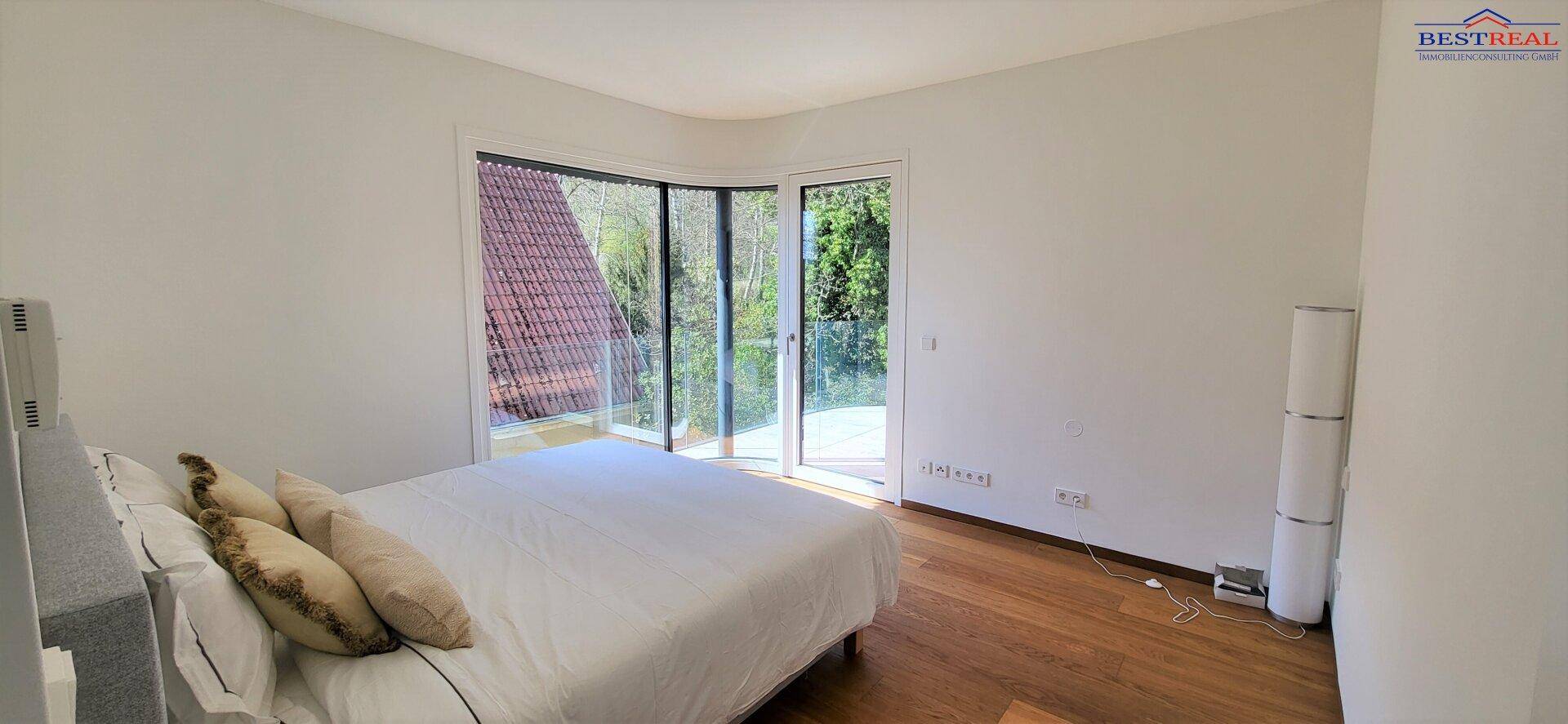Schlafzimmer mit Holzboden und großem Fenster, das Zugang zu einem Balkon bietet und Blick ins Grüne.