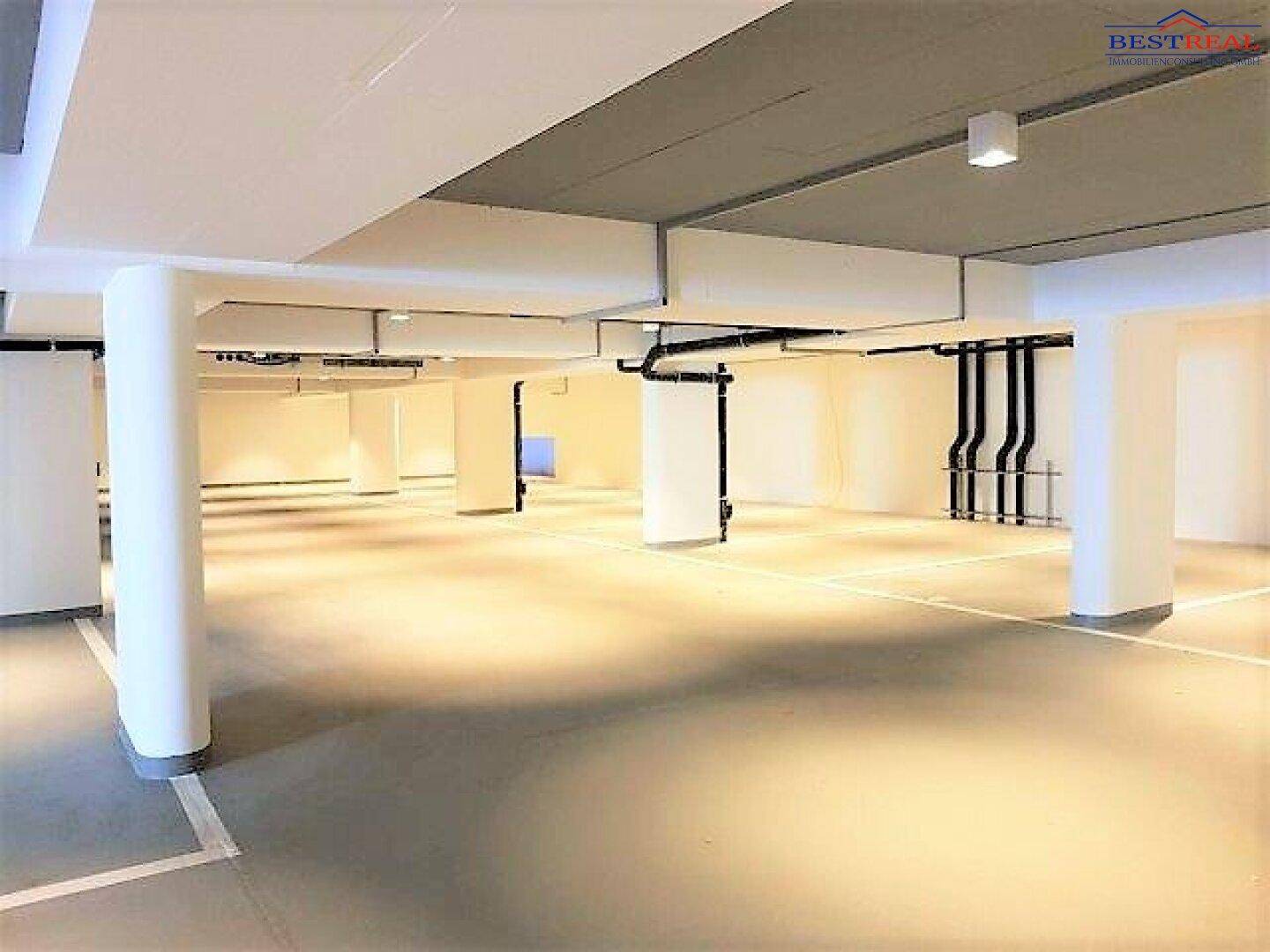 Geräumige Tiefgarage mit markierten Stellplätzen, weißen Säulen und sichtbaren Installationen an der Decke.