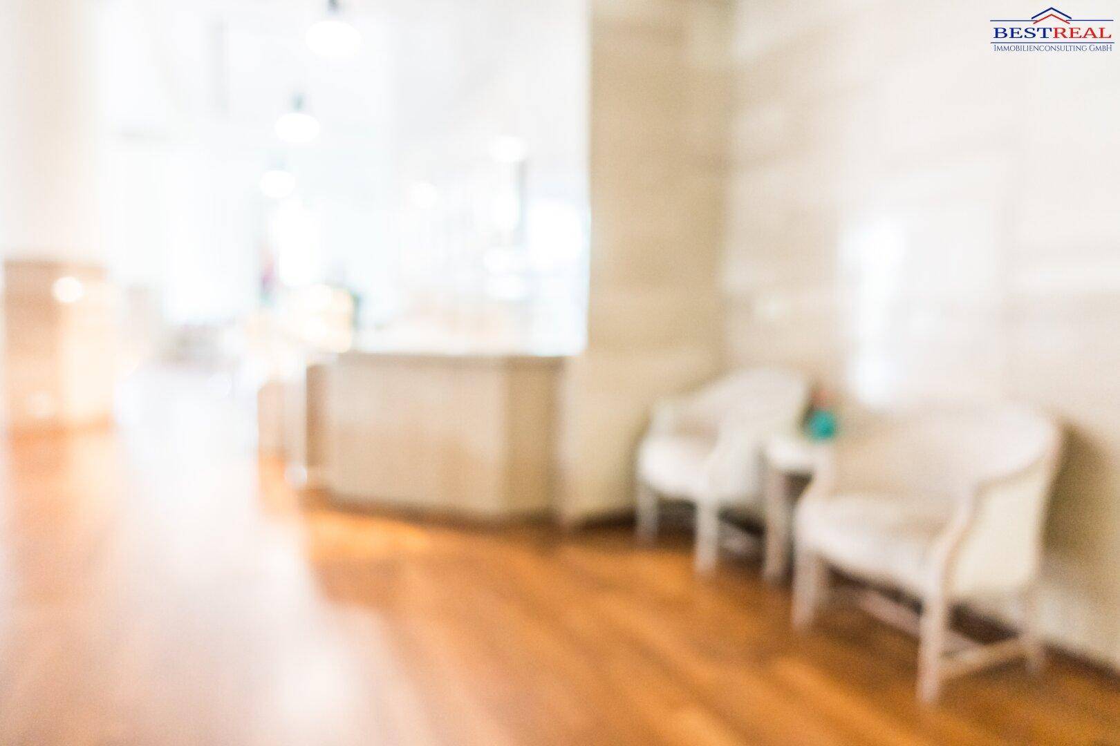 Ein unscharfes Bild eines Innenraums mit Holzboden und hellen Wänden, möglicherweise ein Flur oder Wartebereich.