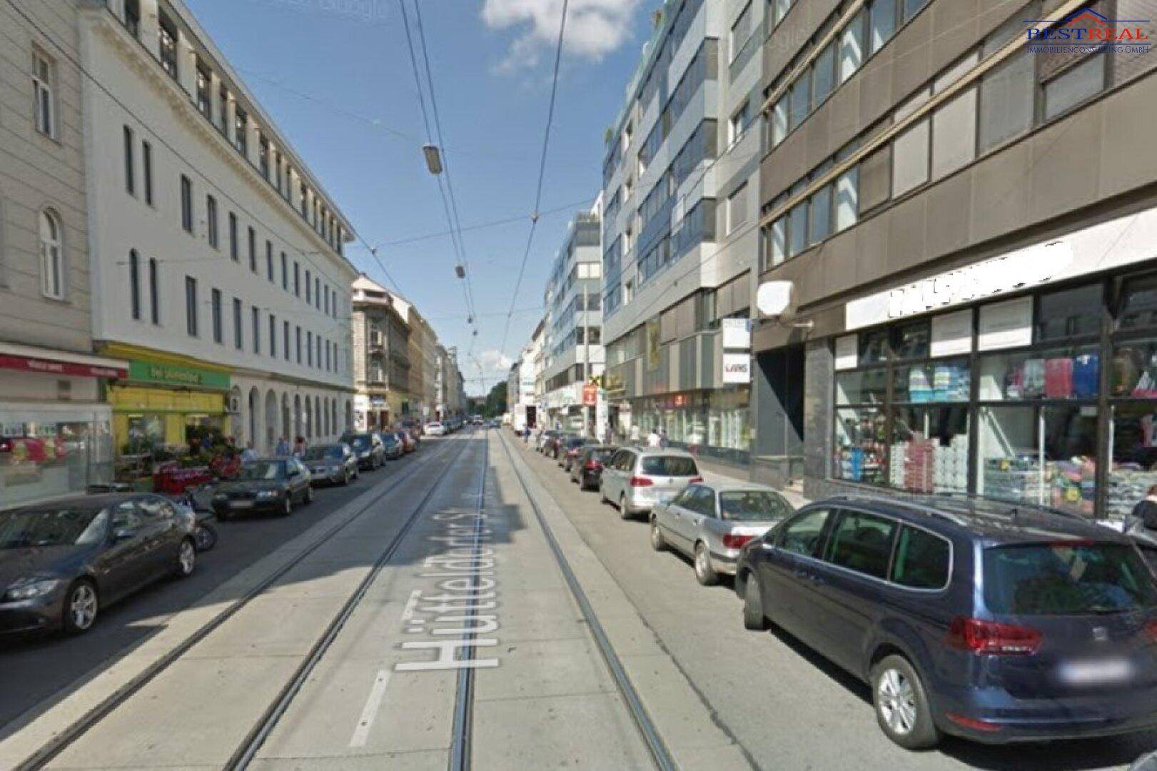 Blick auf eine belebte Stadtstraße mit Gebäuden, geparkten Autos und Straßenbahnschienen unter blauem Himmel.