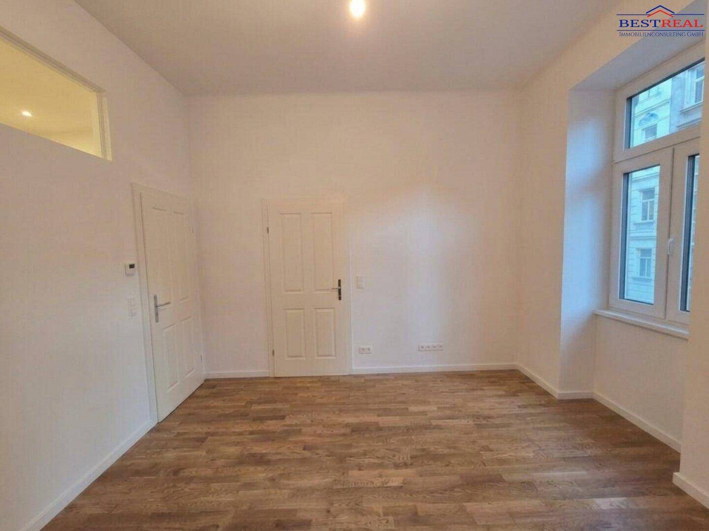 Leeres Zimmer mit Parkettboden, hohen Decken und einem Oberlichtfenster für zusätzliche Helligkeit.