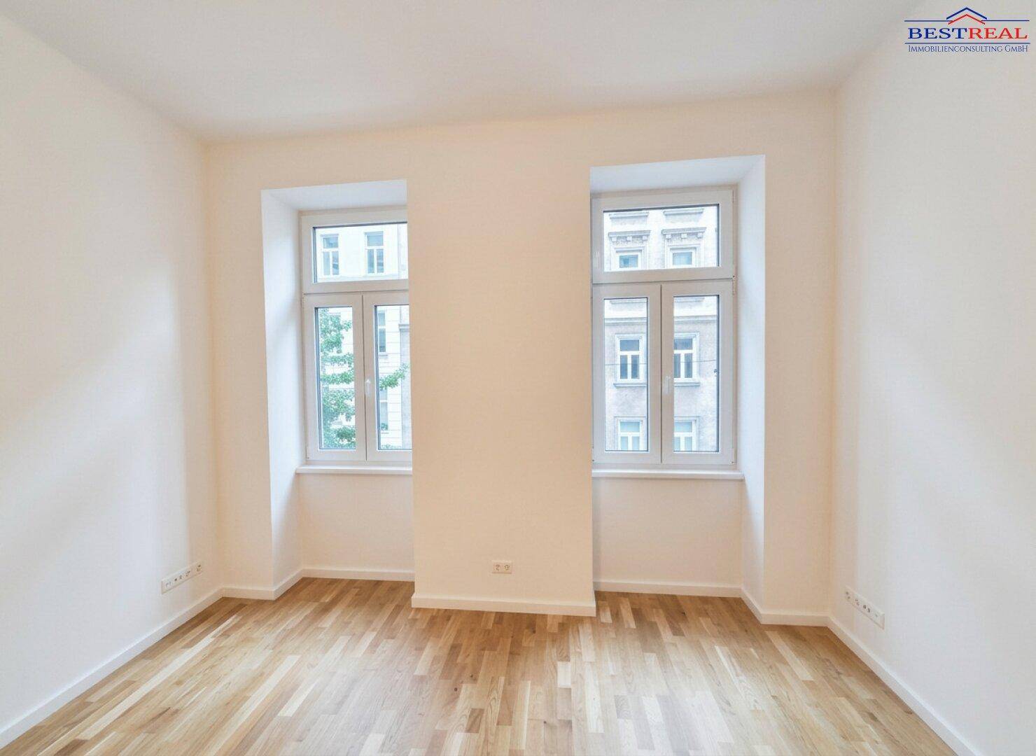 Geräumiges Zimmer mit zwei großen Fenstern und hellem Parkettboden, ideal für Wohn- oder Schlafbereich.
